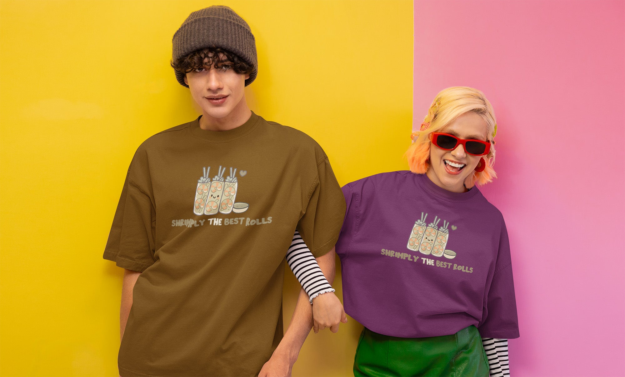 Smiling couple in oversized graphic t-shirts with shrimp spring roll design posing against colorful yellow and pink wall.