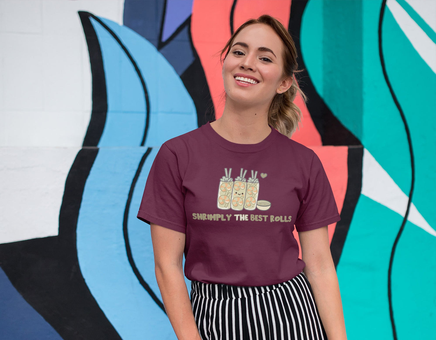 Smiling woman in maroon shrimp spring roll graphic t-shirt posing by colorful abstract mural wall.