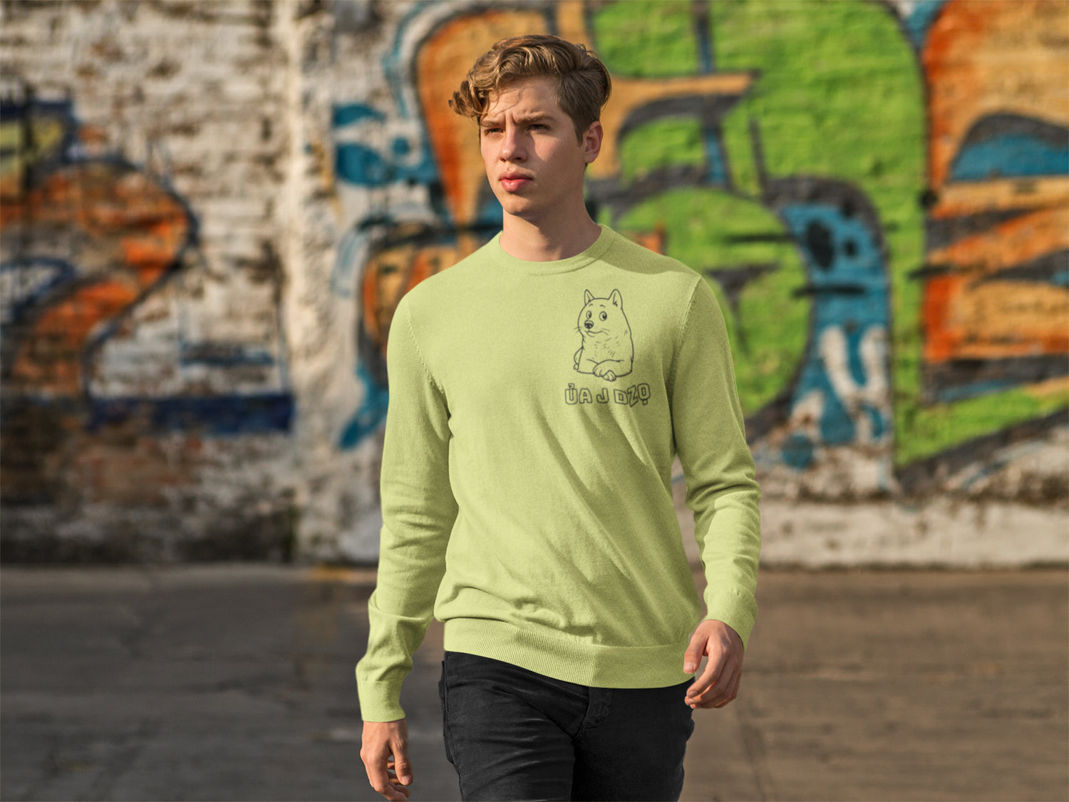 Young man in lime green crewneck with cute dog graphic walking outdoors near graffiti wall.