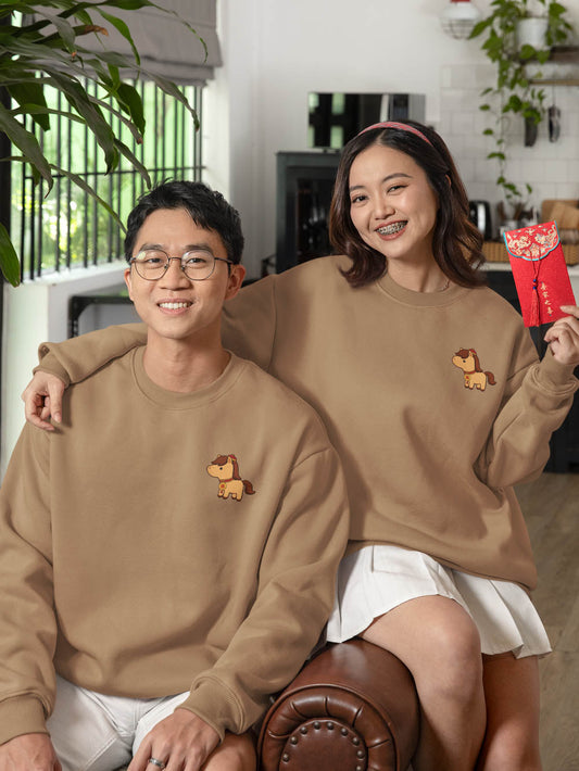 Unisex couple wearing matching crewneck sweatshirts with small embroidered horse design, smiling indoors in a cozy home setting.