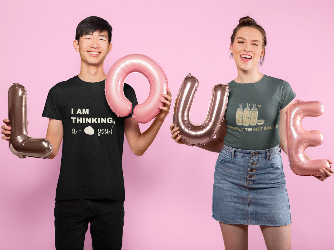 Smiling couple holding pink and gold balloons spelling “love,” wearing cute graphic T-shirts. One says “I Am Thinking, A-Bao You” with a bao bun graphic, and the other says “Shrimply the Best Rolls” with shrimp rolls. 
