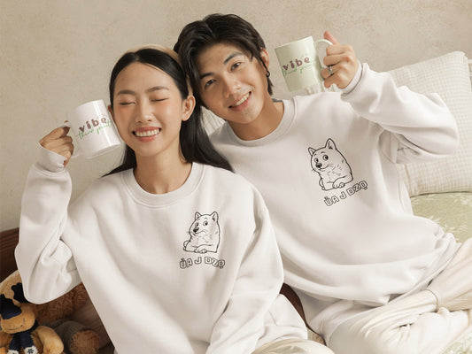 Smiling couple wearing matching white crewnecks with a cute dog illustration and the Vietnamese phrase “Ủa J Dzọ,” holding mugs with “Vibe Thread Print” logo. A cozy, minimalist outfit from a small online apparel brand for teens and young adults.