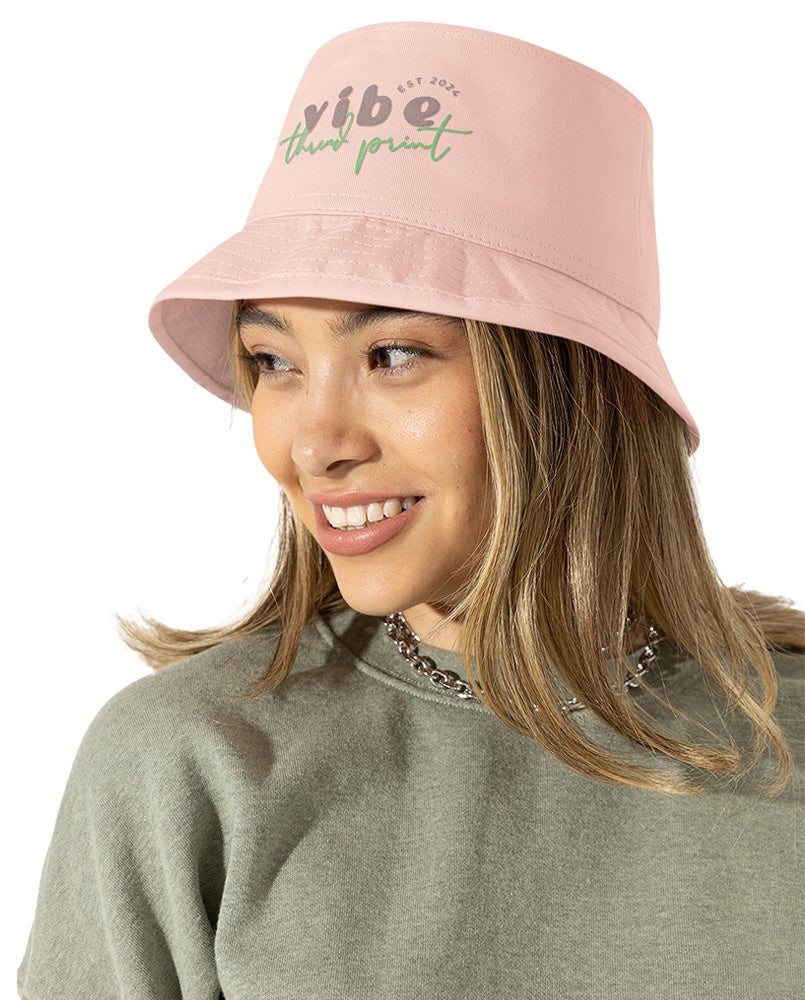 Smiling young woman wearing a pastel pink bucket hat with “Vibe Thread Print” logo embroidery. A cute, minimalist accessory from a small online apparel shop that sells kawaii, cozy streetwear and graphic tees for teens and young adults.