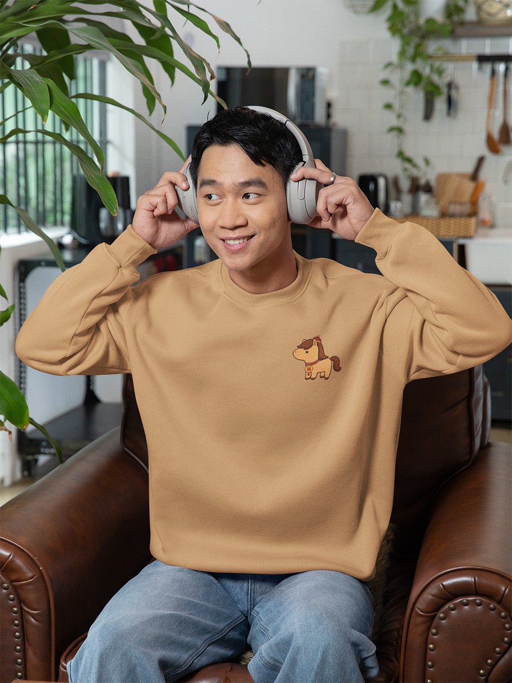 Young man relaxing in khaki oversized crewneck sweatshirt with kawaii horse design, comfy unisex streetwear for teens and young adults.