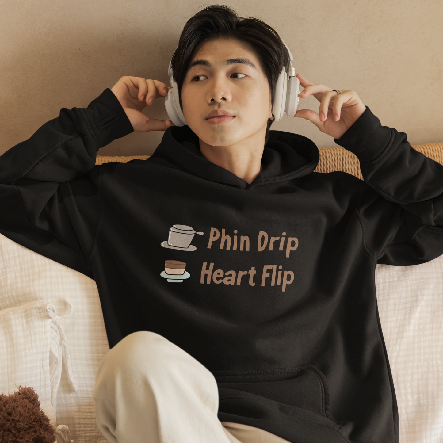 Young man wearing a black graphic hoodie with “Phin Drip Heart Flip” Vietnamese coffee design, relaxing with headphones in cozy casual streetwear for trendy teens and young adults.