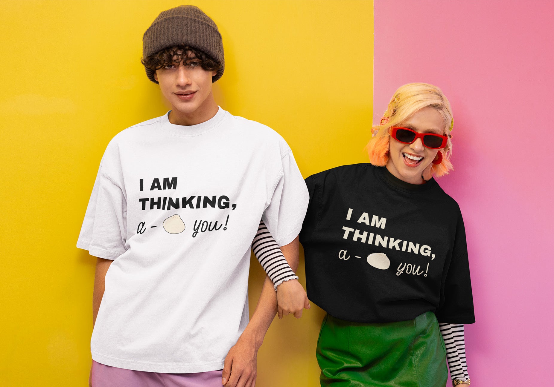 Smiling couple in oversized graphic t-shirts with shrimp spring roll design posing against colorful yellow and pink wall.