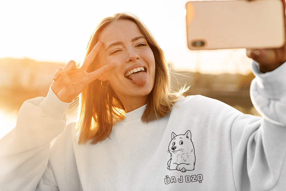 Cheerful young woman taking a selfie in a white crewneck featuring a cute dog illustration and the Vietnamese phrase “Ủa J Dzọ.” Playful, cozy streetwear from a small online apparel shop that sells cute and funny graphic designs for teens and young adults.