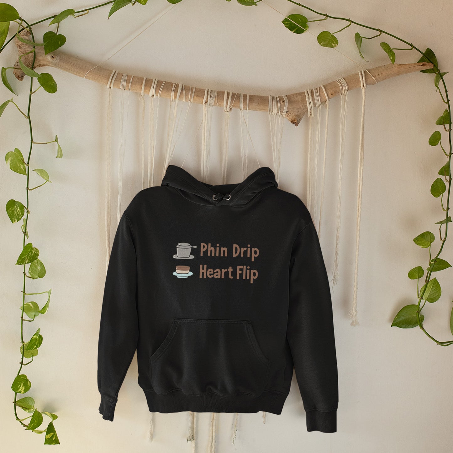 Black hoodie with text and graphics on a white wall with greenery.