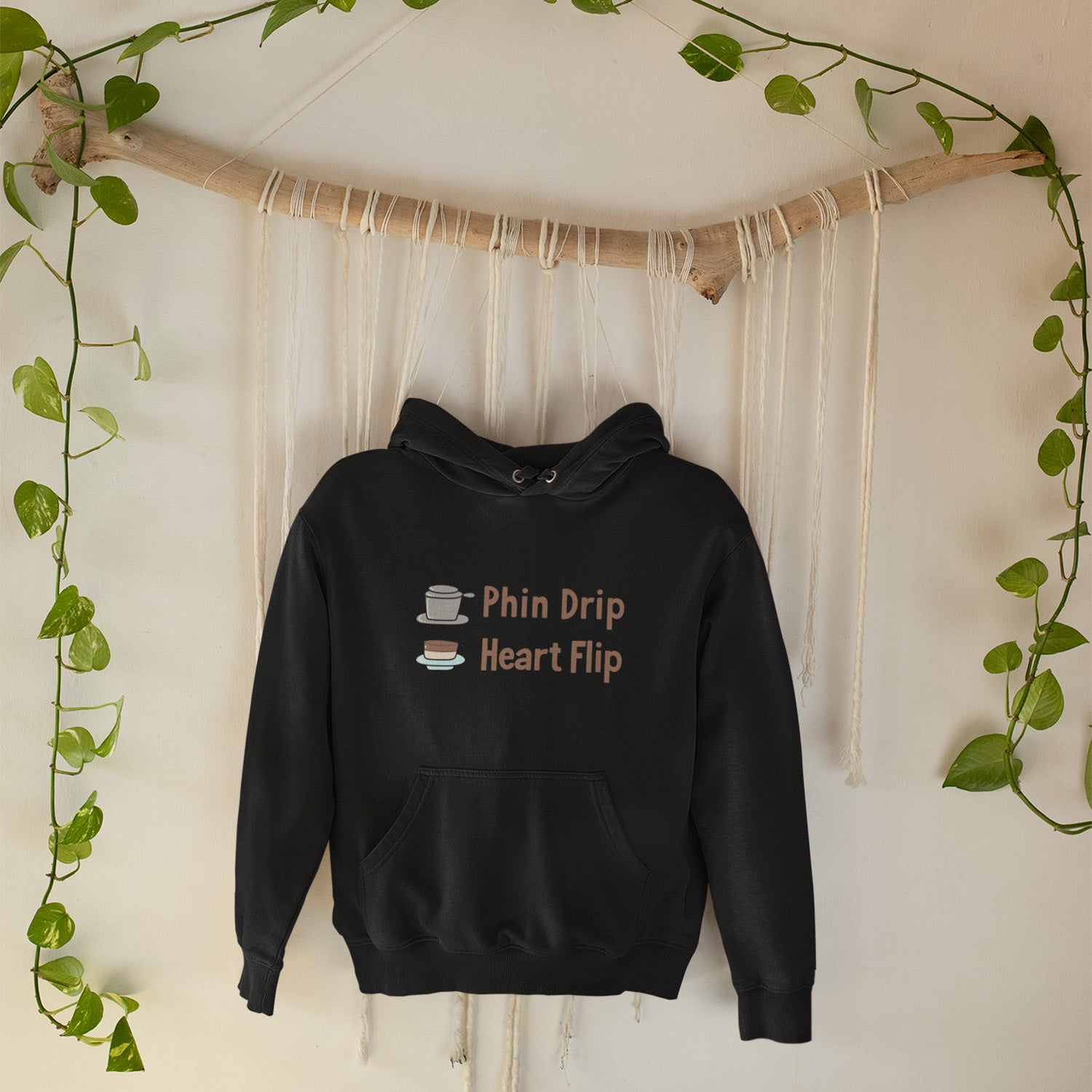 Black hoodie with text and graphics on a white wall with greenery.