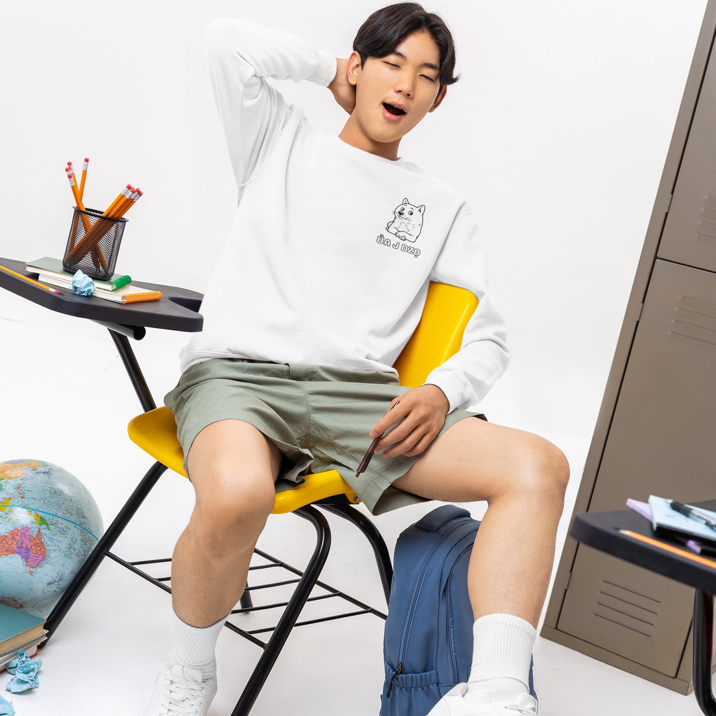 Man yawning in white sweatshirt with dog graphic, sitting in classroom with yellow chair and school supplies.