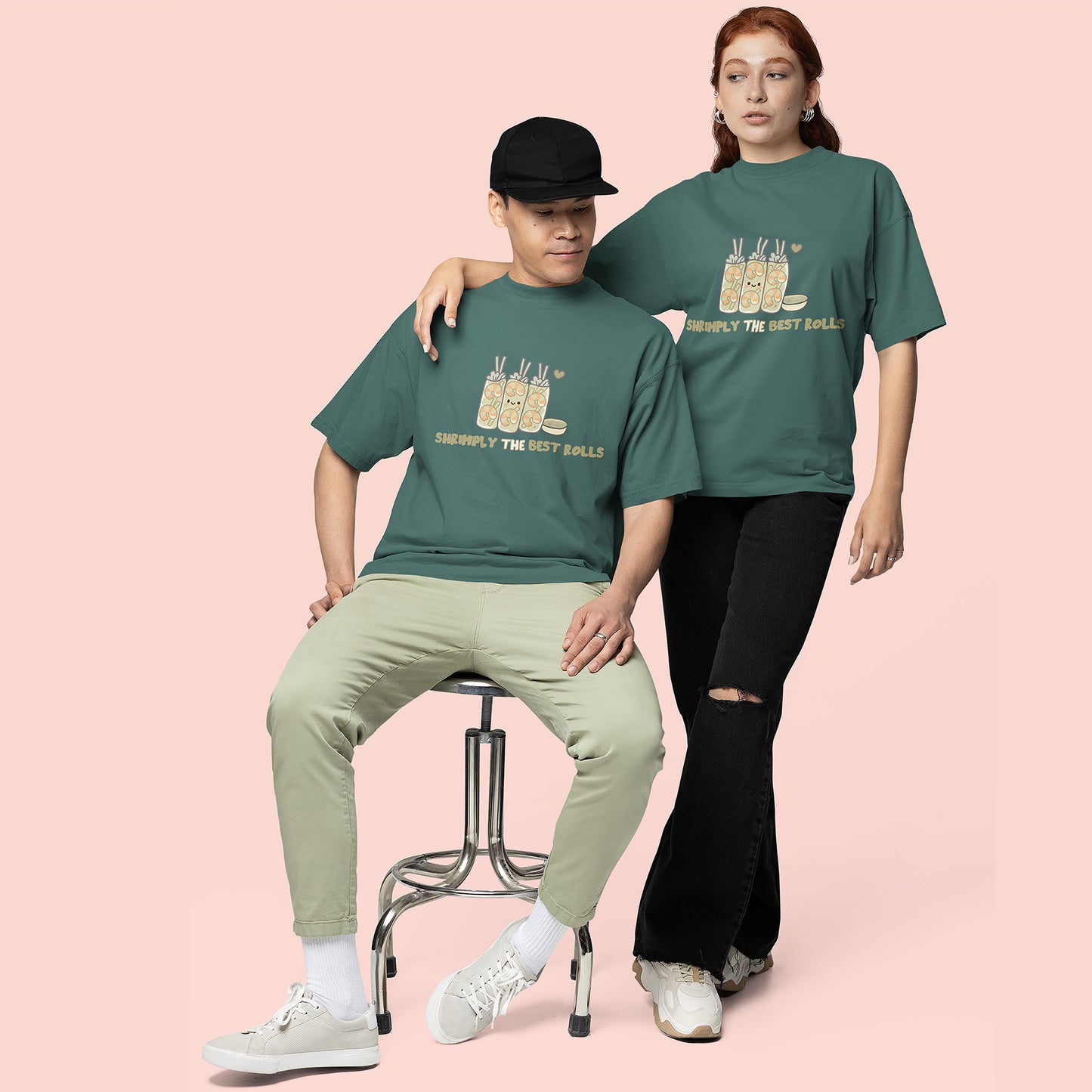 Couple wearing blue spruce “Shrimply the Best Rolls” T-shirts with cute shrimp roll illustration on pastel pink background.